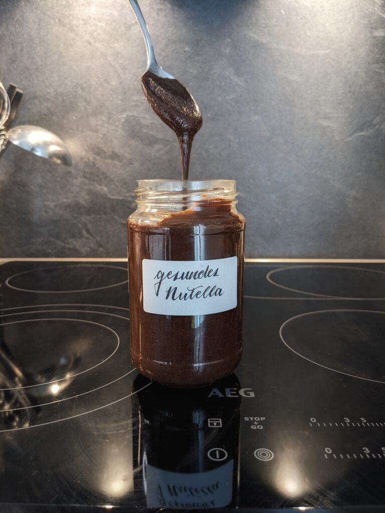 Gesündere Zartbitter Schoko-Haselnuss-Creme in einem Glas, von einem Teelöffel läuft etwas davon zurück ins Glas