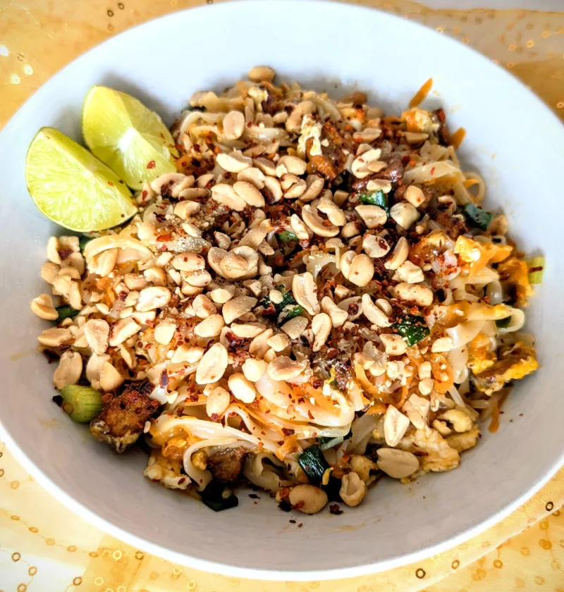 Authentisches Pad Thai mit Reisnudeln, frischem Tofu, Erdnüssen, Limette und Frühlingszwiebeln – auf einem Teller angerichtet, typisch thailändisch und bunt, perfekt für die Follikelphase geeignet. Der Teller ist auf einem weißen und goldfarbigen Tuch angerichtet mit einem holzfarbigen Hintergrund.